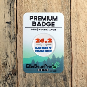 Puede incluir: Una insignia rectangular con esquinas redondeadas, con el texto "PREMIUM BADGE" y "26.2 is my LUCKY NUMBER". La insignia tiene un fondo blanco con una escena de montaña y bosque en la parte inferior. La insignia está sobre una superficie de madera.