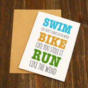 May include: A white card with the text "Swim like there's sharks in the water, Bike like you stole it, Run like the wind" in colorful letters. The card is on a brown background.