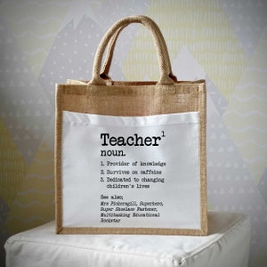 Puede incluir: Una bolsa de lona marrón con un inserto de tela blanca. El inserto tiene la palabra "Teacher" escrita en texto negro, seguida de la definición: "sustantivo. 1. Proveedor de conocimiento 2. Sobrevive a la cafeína 3. Dedicado a cambiar la vida de los niños Ver también; Sra. Pickersgill, Superhéroe, Super sujetador de cordones, Rockstar educativo multitarea"