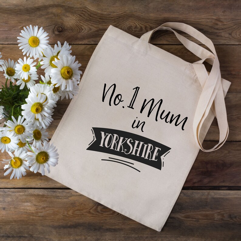 May include: A white canvas tote bag with black text that reads "No. 1 Mum in Yorkshire". The bag has black handles and is sitting on a wooden surface.