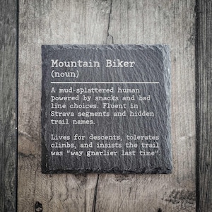 May include: A black slate coaster with the text "Mountain Biker (noun)" and a humorous definition about mountain biking. The coaster is rectangular and sits on a wooden surface.