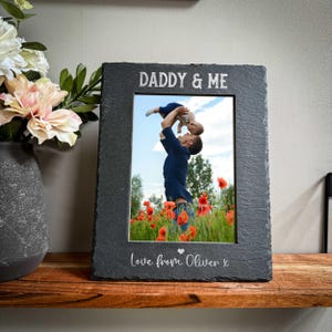 以下が含まれることがあります： 上部に「DADDY & ME」と刻まれた長方形のスレート写真フレーム。 フレームには男性と赤ちゃんの写真が入っています。 フレームの下部には「Love from Oliver x」と書かれています。