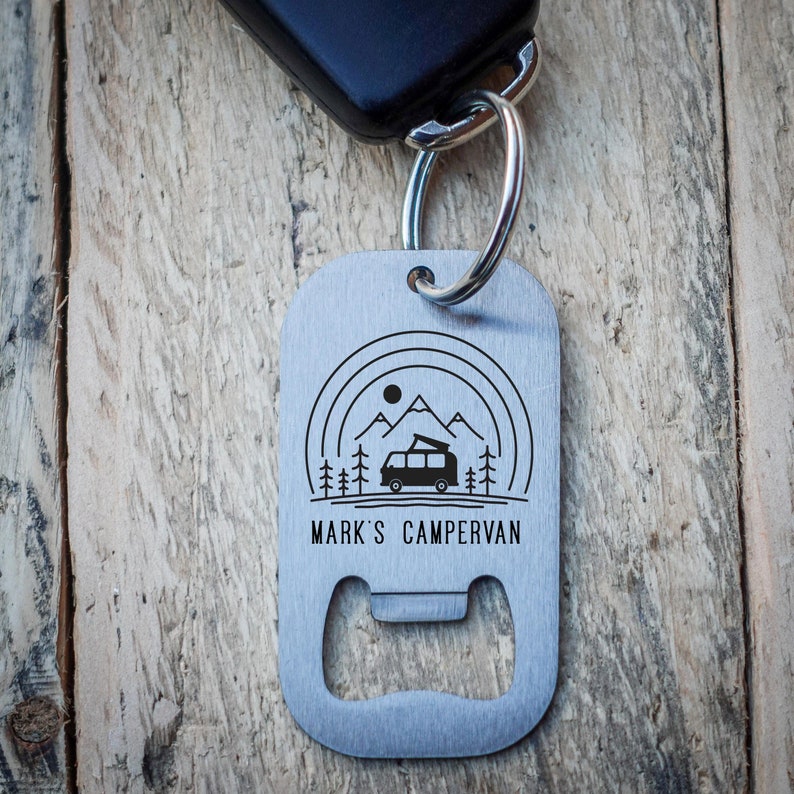 Puede incluir: Un abridor de botellas de llavero plateado con un dise&ntilde;o en blanco y negro de una autocaravana frente a monta&ntilde;as. El llavero tambi&eacute;n tiene el texto "Mark's Campervan" grabado.