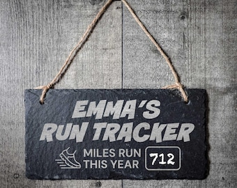 Może przedstawiać: Prostokątny czarny znak z łupków z białym napisem "EMMA'S RUN TRACKER". Poniżej napis "MILES RUN THIS YEAR" z grafiką buta do biegania i numerem "712". Znak zawieszony na sznurku jutowym.