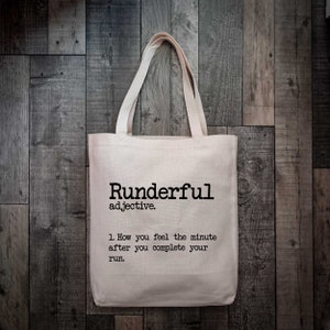 Puede incluir: Una bolsa de tela de color beige con texto impreso en negro que dice: Runderful adjective. 1. How you feel the minute after you complete your run. La bolsa tiene dos asas de tela a juego y una forma rectangular con una base plana.