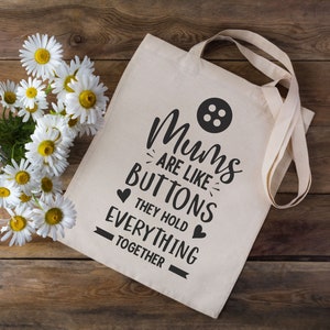 May include: A white canvas tote bag with black text that reads "Mums are like buttons they hold everything together". The bag has a black button graphic and two black hearts.