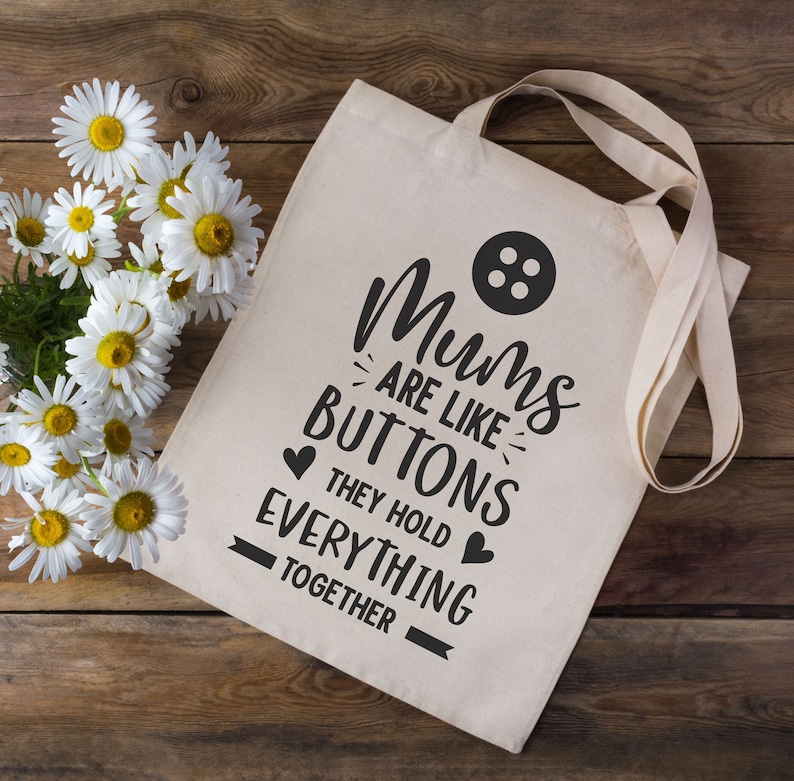 May include: A white canvas tote bag with black text that reads "Mums are like buttons they hold everything together". The bag has a black button graphic and two black hearts.