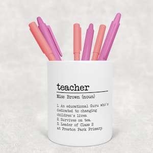 Puede incluir: Porta lápices de cerámica blanca con la palabra "teacher" y el texto "Miss Brown (noun)" seguido de una definición. El porta lápices tiene cinco bolígrafos rosas dentro.