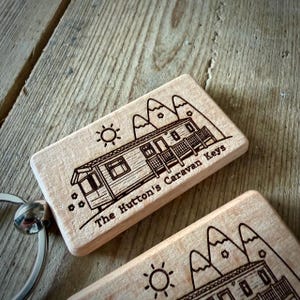 May include: A rectangular light-coloured wooden keyring featuring a laser-engraved illustration of a caravan, mountains, and a sun. The text "The Hutton's Caravan Keys" is engraved on the surface. A silver metal split ring is attached to the side.