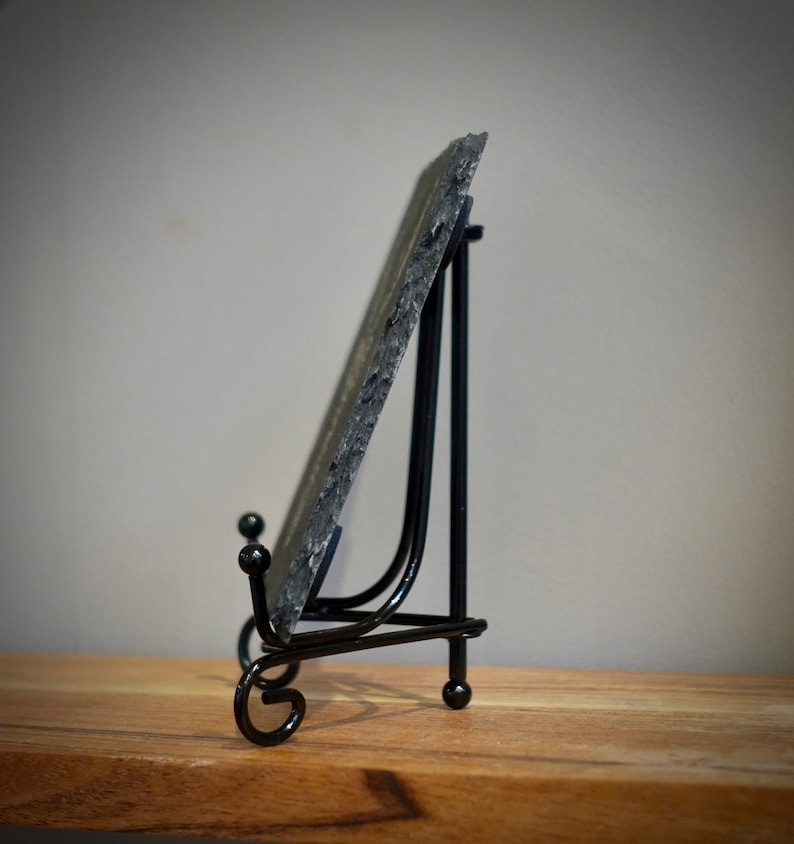 Peut inclure: Un chevalet en m&eacute;tal noir avec une plaque en ardoise. Le chevalet a un motif de volutes d&eacute;coratif &agrave; la base.