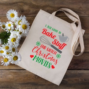 May include: A cream-colored tote bag with the text "I just want to bake stuff and watch Christmas movies" in red and green. The design includes a whisk, measuring cup, snowflakes, and hearts. The bag has long straps.