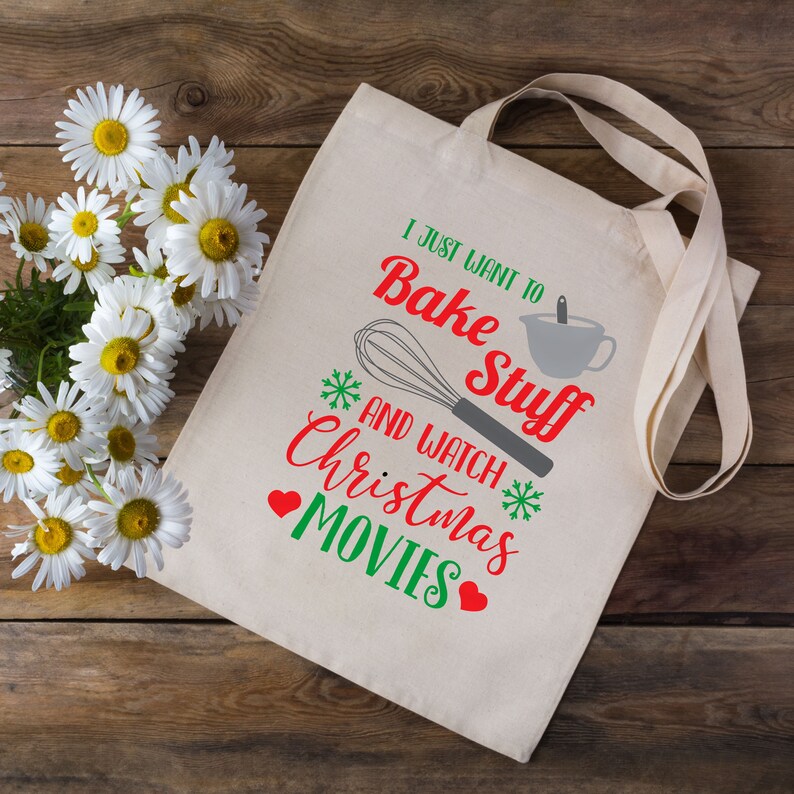 May include: A cream-colored tote bag with the text "I just want to bake stuff and watch Christmas movies" in red and green. The design includes a whisk, measuring cup, snowflakes, and hearts. The bag has long straps.