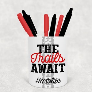 May include: White ceramic pencil holder with a black and grey tyre tread design. The text "THE Trails AWAIT #mtblife" is printed on the holder in black and red.