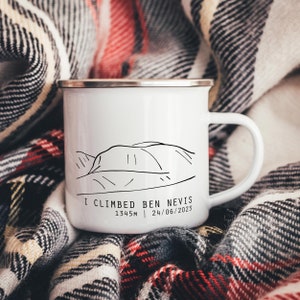 Può includere: Tazza di smalto bianco con un disegno a linea nera di una catena montuosa e il testo "I climbed Ben Nevis 1345m | 24/06/2023". La tazza è appoggiata su una coperta a quadri.