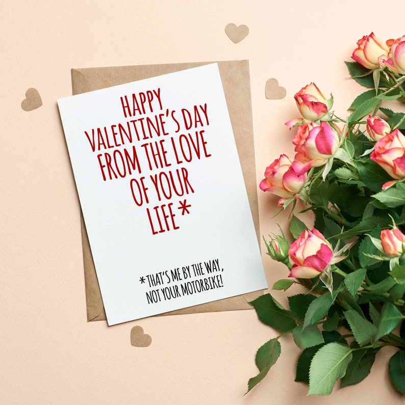 K&ouml;nnte beinhalten: Eine wei&szlig;e Karte mit rotem Text, der "Happy Valentine's Day from the love of your life* *That's me by the way, not your motorbike!" lautet. Die Karte ist von rosa Rosen und braunen Papierherzen umgeben.