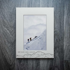 May include: White framed print with a photograph of two climbers ascending a snowy mountain. The print includes the text "MONT BLANC | 4809m | 19/06/2024" and a black line drawing of mountains at the bottom.