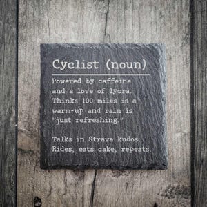 以下が含まれることがあります： 「Cyclist (noun)」のテキストと、サイクリストの習慣と好みをユーモラスに表現した、正方形の濃い灰色のスレートコースター。コースターは木製の背景に置かれています。