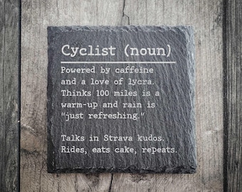 May include: A square, dark gray slate coaster with the text "Cyclist (noun)" and a humorous description of a cyclist's habits and preferences. The coaster is set against a wooden background.
