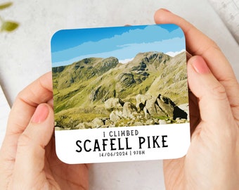 May include: A white coaster with a colourful image of a mountain range and the text "I climbed Scafell Pike 14/06/2024 | 978m".