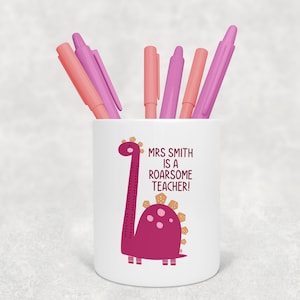 May include: White ceramic pencil holder with a pink dinosaur design. The dinosaur has a long neck and a gold crown. The text on the holder reads "Mrs. Smith is a roarsome teacher!"