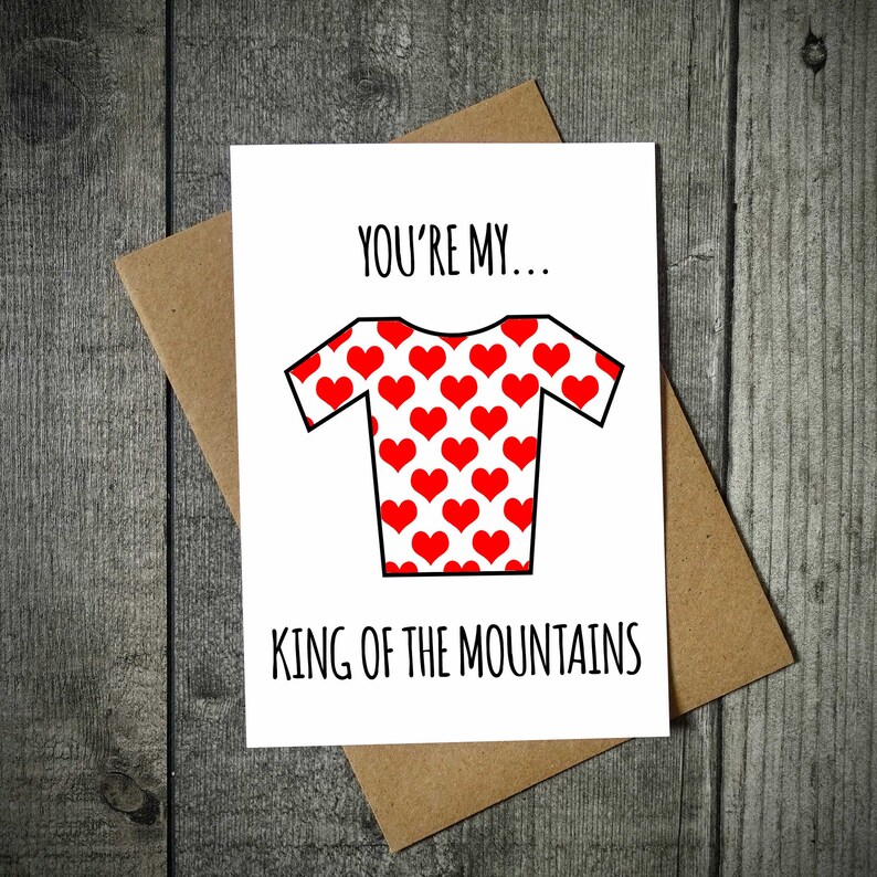 May include: A white greeting card with the text "YOU'RE MY... KING OF THE MOUNTAINS" and a red heart-patterned jersey design. The card is placed on a brown envelope, set against a wooden background.
