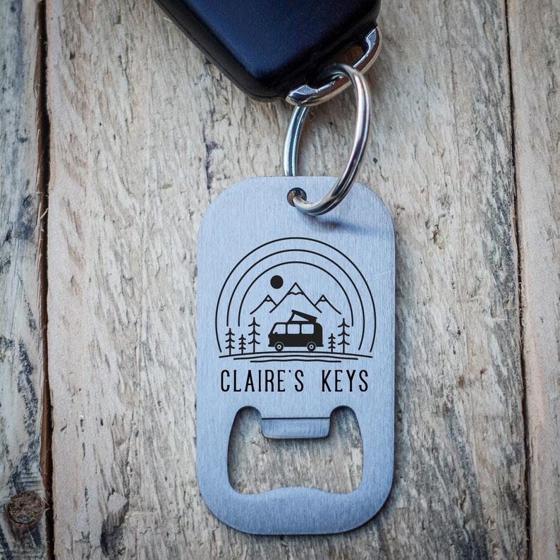 Puede incluir: Un llavero de metal plateado con un abridor de botellas. El llavero presenta un gr&aacute;fico de una autocaravana conduciendo por un paisaje monta&ntilde;oso con &aacute;rboles y un sol. El texto "CLAIRE'S KEYS" est&aacute; grabado en el llavero.