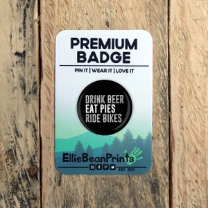 May include: Black and white button badge with the text "Drink Beer Eat Pies Ride Bikes". The badge is on a white card with the text "Premium Badge Pin It | Wear It | Love It" and the text "Ellie Bean Prints EST. 2011".