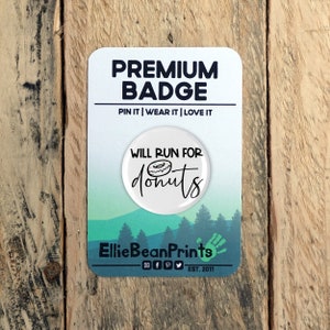 Könnte beinhalten: Runder weißer Anstecker mit schwarzem Text "Will Run For Donuts" und einer schwarz-weißen Donut-Illustration. Der Anstecker befindet sich auf einer weißen Karte mit grünem und blauem Hintergrund und dem Text "Premium Badge" und "Pin It | Wear It | Love It". Die Karte enthält außerdem den Text "Ellie Bean Prints" mit Social-Media-Symbolen und "EST. 2011".
