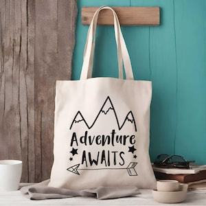 May include: A white canvas tote bag with black text that reads "Adventure Awaits" and a mountain range graphic. The bag also features a black arrow with stars on either side.