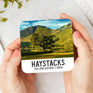 Puede incluir: Una ilustración colorida de la montaña Haystacks en el Lake District, Inglaterra. La imagen muestra una montaña verde con un lago en primer plano y un gran árbol en el medio. El texto "HAYSTACKS THE LAKE DISTRICT | 597m" está impreso debajo de la imagen.