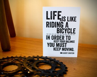 May include: A white card with the quote "Life is like riding a bicycle. In order to keep your balance, you must keep moving." Black bicycle gears are in the foreground. The quote is attributed to Albert Einstein.