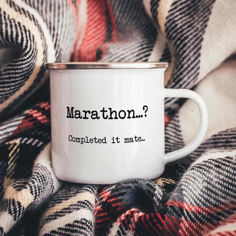 K&ouml;nnte beinhalten: Wei&szlig;er Emaillebecher mit silbernem Rand. Der Becher tr&auml;gt den schwarzen Text "Marathon...? Completed it mate...". Der Becher steht auf einer karierten Decke mit roten, wei&szlig;en und schwarzen Mustern.