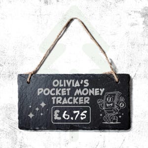 May include: A black slate pocket money tracker sign with the text "OLIVIA'S POCKET MONEY TRACKER" and a cartoon money character. The sign has a rope hanger and the amount £6.75 is displayed.