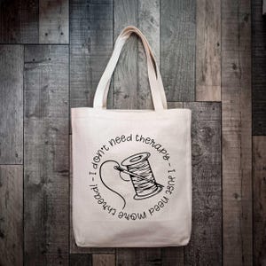 May include: A beige canvas tote bag featuring a black graphic of a spool of thread with a needle. The text surrounding the illustration reads, I don't need therapy, I just need more thread. The bag has two shoulder straps and a rectangular shape.