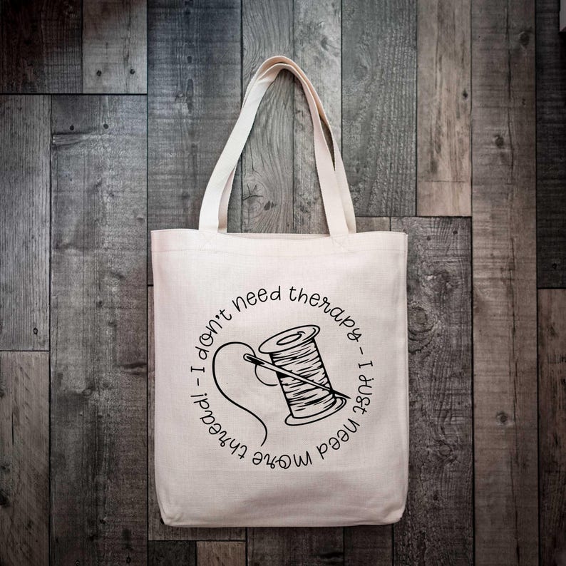 May include: A beige canvas tote bag featuring a black graphic of a spool of thread with a needle. The text surrounding the illustration reads, I don't need therapy, I just need more thread. The bag has two shoulder straps and a rectangular shape.