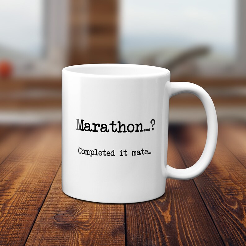 K&ouml;nnte beinhalten: Wei&szlig;e Keramik-Tasse mit dem Text "Marathon...? Completed it mate..." in Schwarz. Die Tasse hat einen gebogenen Henkel und steht vor einem verschwommenen Hintergrund auf einer Holzoberfl&auml;che.