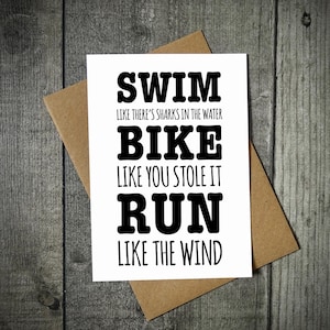 Peut inclure: Une carte de vœux rectangulaire blanche avec un texte noir qui indique : SWIM LIKE THERE'S SHARKS IN THE WATER, BIKE LIKE YOU STOLE IT, RUN LIKE THE WIND. La carte est placée sur une enveloppe en papier kraft brun, créant un contraste visuel.