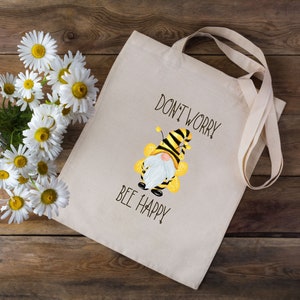 May include: Beige tote bag with the text "DON'T WORRY BEE HAPPY" and a cartoon bee gnome illustration. The bag has long handles and is next to a bouquet of white daisies with yellow centers.