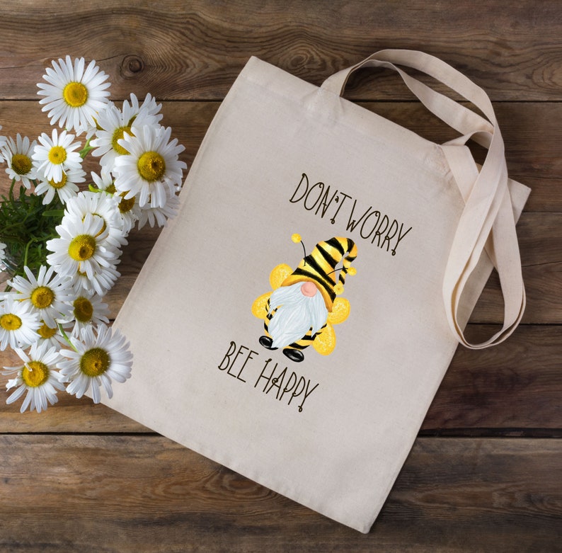 May include: Beige tote bag with the text "DON'T WORRY BEE HAPPY" and a cartoon bee gnome illustration. The bag has long handles and is next to a bouquet of white daisies with yellow centers.
