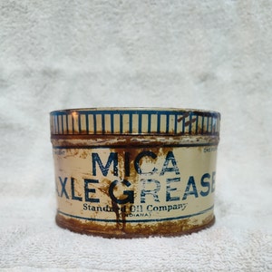 May include: A vintage tin can of Mica Axle Grease, with blue and white striped lettering on a cream background. The text reads "Mica Axle Grease" and "Standard Oil Company (Indiana)".