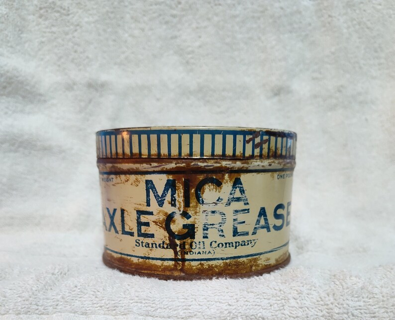 May include: A vintage tin can of Mica Axle Grease, with blue and white striped lettering on a cream background. The text reads "Mica Axle Grease" and "Standard Oil Company (Indiana)".