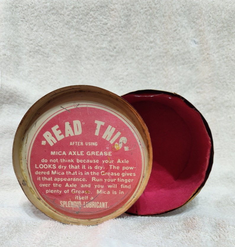 May include: A red and white tin container with the text "READ THIS" and "MICA AXLE GREASE" on the lid. The text reads "do not think because your Axle LOOKS dry that it is dry. The powdered Mica that is in the Grease gives it that appearance. Run your finger over the Axle and you will find plenty of Grease. Mica is in itself a SPLENDID LUBRICANT."