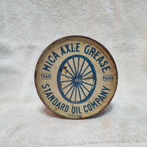 May include: A round, white metal container with a blue and white label. The label reads "MICA AXLE GREASE TRADE MARK STANDARD OIL COMPANY (INDIANA)" and features a blue wheel with spokes.