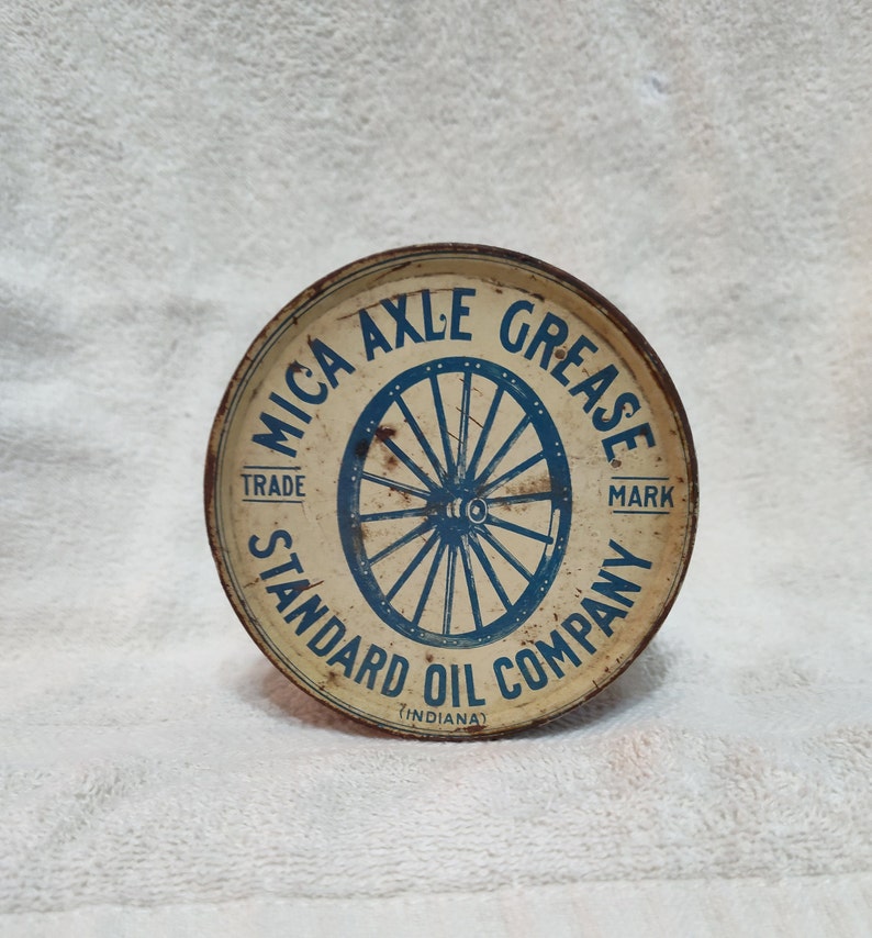 May include: A round, white metal container with a blue and white label. The label reads "MICA AXLE GREASE TRADE MARK STANDARD OIL COMPANY (INDIANA)" and features a blue wheel with spokes.