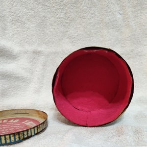 May include: An empty, round, vintage tin container with a pink interior. The lid is off and shows the text "Bread  Twis" with a graphic of a braid of bread.