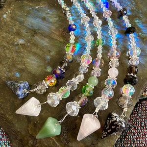 May include: Five crystal pendulums with different colored stones at the bottom. The pendulums are hanging from a clear crystal beaded chain. The stones are green, pink, black, blue, and a multi-colored stone.