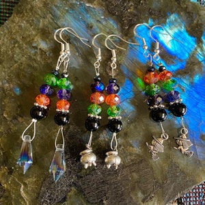 May include: Three pairs of Halloween-themed dangle earrings. Each pair features a combination of colorful beads in orange, green, blue, and black. One pair has crystal pendants, another has cauldron charms, and the last has witch charms.