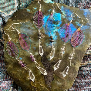 May include: Three pairs of iridescent leaf-shaped earrings with crystal and gemstone accents. The earrings are displayed on a dark gray stone.