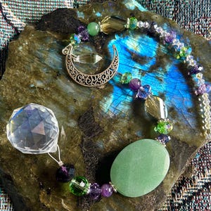 May include: A crystal pendant necklace with a faceted crystal ball, a green oval stone, and a silver crescent moon charm. The necklace features green, purple, and clear beads. The necklace is displayed on a dark, textured surface.