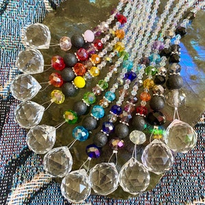 May include: A collection of crystal suncatchers with rainbow coloured beads and black lava rock beads. The suncatchers are hanging from a light grey surface.